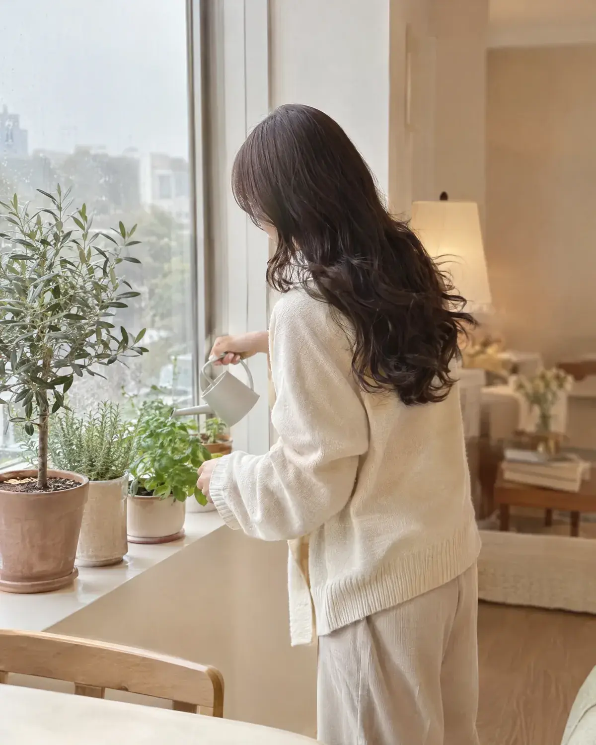 윗머리는 차분하고 아래 층에 컬이 있는 긴 머리 여성이 창가 화분에 물을 주는 뒷모습