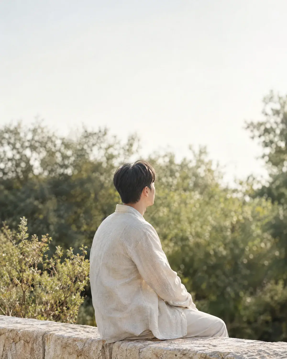 짧게 정리한 머리의 남성이 돌담에 앉아 나무를 바라보는 뒷모습