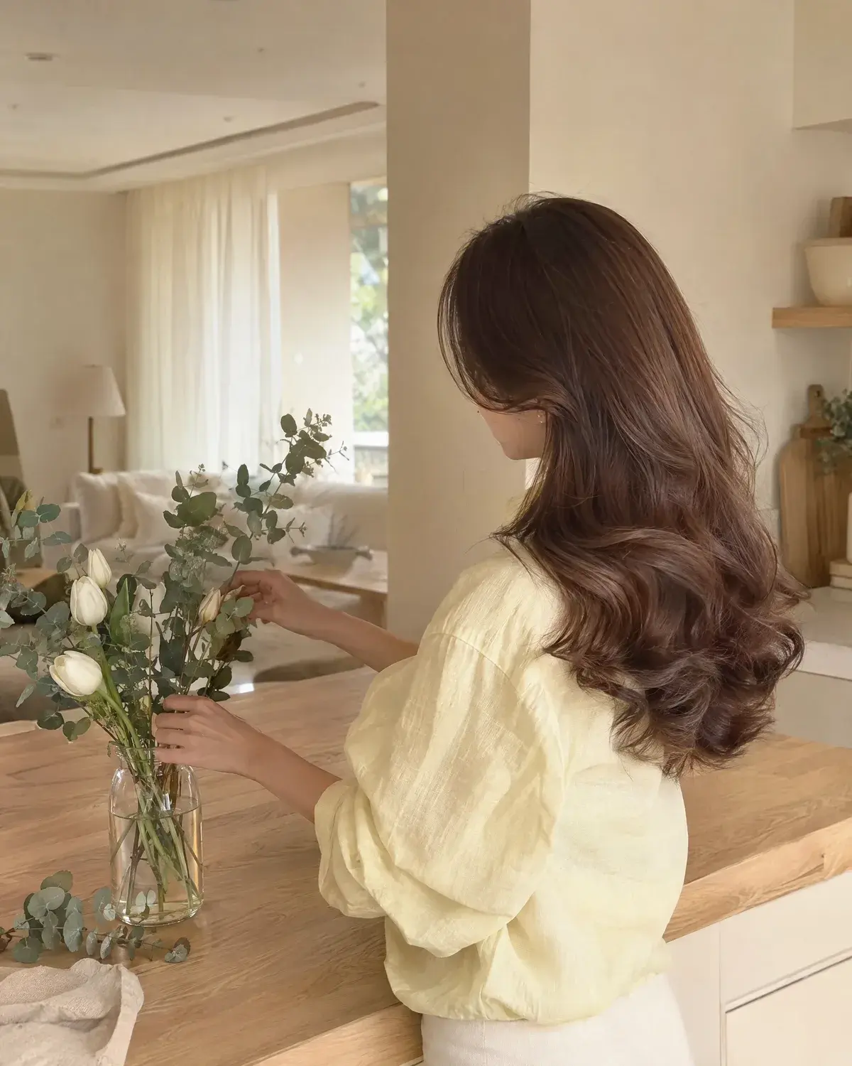 중간부터 둥글고 풍성한 컬이 들어간 긴 밤색 머리 여성이 주방에서 튤립을 꽂는 옆뒷모습