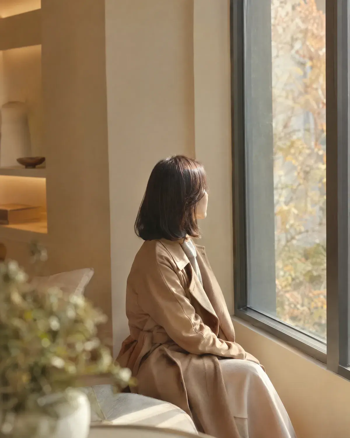 트렌치코트를 입은 단발 여성이 창가 벤치에 앉아 창밖을 바라보는 옆모습